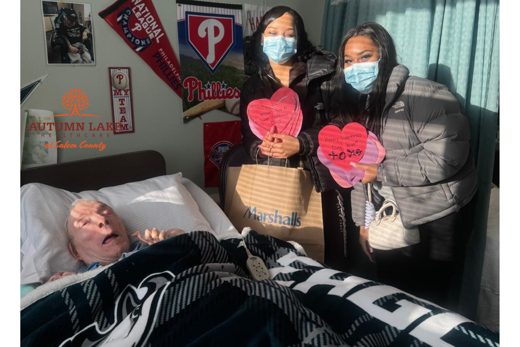 Two masked visitors stand by a resident's bed in an assisted living room, holding handmade Valentine's hearts and a gift bag.