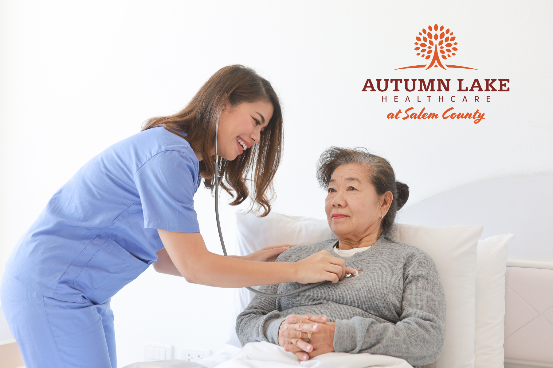 A smiling nurse in blue scrubs uses a stethoscope to check the vitals of an elderly woman resting in bed at Autumn Lake Healthcare at Salem County.